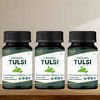 Three bottles of Organic Tulsi dietary supplements on a wooden surface with a beige background.